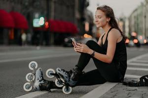 photo horizontale d'une femme sportive active vêtue de vêtements de sport porte des rollers concentrés sur un smartphone aime le patin à roues alignées à l'extérieur mène un style de vie actif. roller comme concept de passe-temps