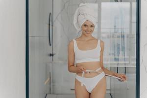 bonne fille en forme positive posant dans la salle de bain après la douche du matin avec un ruban à mesurer tenant autour de sa taille photo