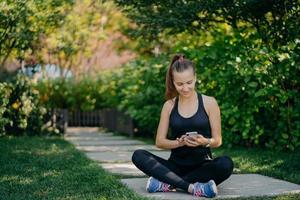 photo en extérieur d'une femme athlétique se repose après l'entraînement s'assoit les jambes croisées dans le parc surfe sur les réseaux sociaux vêtu de vêtements de sport écoute de la musique via des écouteurs sans fil utilise l'application de sport bénéficie du beau temps