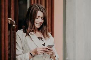 jeune femme souriante aux cheveux noirs, vêtue d'une élégante veste blanche, tient un smartphone moderne, se tient près des portes d'un immeuble de bureaux, envoie des messages à des amis sur les réseaux sociaux. notion de technologie photo