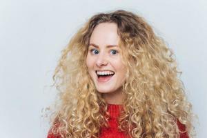 photo de tête d'une belle femme satisfaite avec une expression joyeuse agréable, avec des cheveux bouclés et touffus, vêtue d'un pull rouge, ne s'attend pas à entendre quelque chose d'excité, isolée sur fond blanc