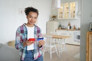 une fille afro-américaine heureuse achète en ligne avec un téléphone portable et une carte de crédit depuis chez elle. photo