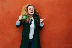 femme excitée aux cheveux blonds bouclés portant une veste tenant une tasse de café verte levant son pouce montrant sa satisfaction à boire avec un large sourire. jolie femme appréciant de boire du café photo