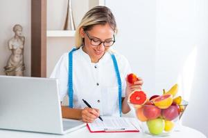 nutritionniste souriante dans son bureau, elle tient un fruit et montre des légumes et des fruits sains, des soins de santé et un concept de régime. femme nutritionniste avec des fruits travaillant à son bureau photo