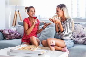 deux jolies filles, de joyeuses meilleures amies s'amusant, buvant du vin rouge et mangeant de la pizza à la maison. verre de vin et tranche de pizza dans les mains. les filles s'amusent ensemble à bavarder photo