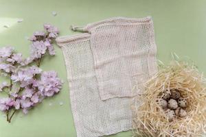 branches de fleurs de cerisier, sacs de fruits en textile, fleurs roses, nid d'oiseau avec des œufs de caille sur fond texturé vert clair, vacances de printemps concept écologique festif, flaylay, espace de copie pour le texte photo