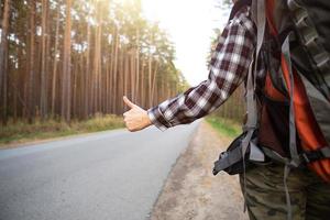 main avec les pouces vers le haut sur le fond de l'autoroute - auto-stop, vote. touriste dans une chemise à carreaux avec sac à dos près de la forêt faire du stop. auto-stop, tourisme intérieur, aventure seul, voyage, randonnée photo