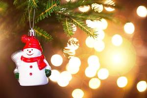 jouet de noël bonhomme de neige dans un chapeau rouge avec une écharpe sur la branche vivante d'un sapin avec des lumières dorées de guirlandes en défocalisation. nouvel an, noël, fond de vacances, bokeh, espace de copie photo