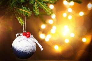 boule de noël avec de la neige, des baies rouges et un arc sur une branche vivante d'un sapin avec des lumières dorées de guirlandes en défocalisation. nouvel an, noël, fond de vacances, bokeh, espace de copie photo