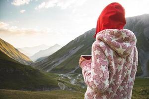 vue arrière femme caucasienne réfléchie en pyjama debout sur le point de vue boire du café frais dans la nature profiter du panorama pittoresque des montagnes au lever du soleil tôt le matin seul. photo