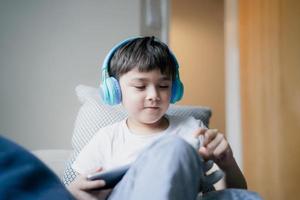 heureux jeune garçon portant un casque pour jouer à un jeu sur internet avec des amis, enfant assis à côté de la fenêtre lisant ou regardant un dessin animé sur une tablette, écolier faisant ses devoirs en ligne à la maison. photo
