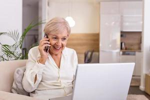 femme âgée travaillant sur un ordinateur portable, souriant, parlant au téléphone. femme âgée utilisant un ordinateur portable. femme âgée assise à la maison, utilisant un ordinateur portable et parlant sur son téléphone portable, souriante. photo