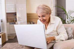 portrait d'une femme âgée assise à table à la maison et travaillant sur son ordinateur portable. dame âgée surfant sur le net depuis la maison assise sur son canapé et utilisant un ordinateur portable photo