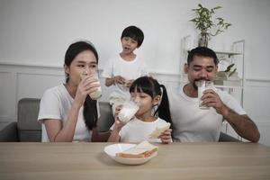 une famille thaïlandaise asiatique en bonne santé, de petits enfants et de jeunes parents boivent du lait blanc frais dans du verre et du pain joie ensemble à une table à manger le matin, bien-être nutrition maison petit déjeuner repas style de vie. photo