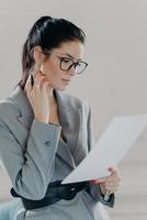 une directrice exécutive réussie parcourt des documents papier, analyse des données, étudie des informations sur les contrats, tient un stylo, porte des lunettes, une tenue formelle, examine le rapport mensuel ou le curriculum vitae des employés photo