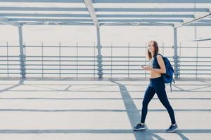 photo en extérieur d'une belle femme au corps mince, vêtue d'un haut et de leggings décontractés, porte un sac, va à la salle de sport, utilise un smartphone moderne pour écouter de la musique, copiez l'espace de côté pour votre publicité