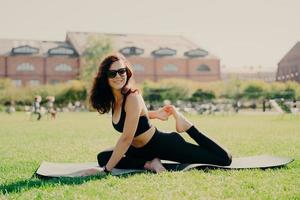 concept d'activité sportive. photo horizontale d'une femme heureuse et motivée qui s'étend sur des trains de tapis de fitness à l'extérieur pendant la journée d'été a un corps sain et sportif porte des lunettes de soleil haut court et des leggings