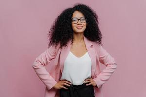 photo à la taille d'une employée de bureau satisfaite à la peau foncée qui garde les mains sur sa silhouette mince, a un regard heureux, porte des lunettes transparentes, pose sur fond violet, prête à parler avec un collègue