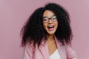 portrait d'une jeune femme aux cheveux bouclés ravie se sent trop émotive, garde la bouche largement ouverte, porte des lunettes optiques et un costume formel, rit joyeusement aux nouvelles, pose à l'intérieur sur fond clair photo