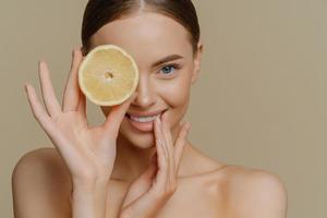 photo intérieure d'un joli modèle féminin a une peau fraîche et saine un corps bien soigné couvre les yeux avec une tranche d'orange pose des épaules nues isolées sur fond beige. ingrédient pour faire un masque facial