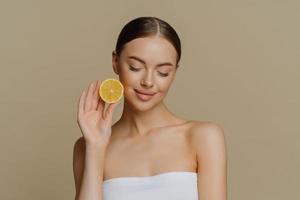 portrait d'une jolie jeune femme brune concentrée tient une tranche de citron frais recommande des produits de beauté naturels préfère un traitement facial biologique naturel enveloppé dans une serviette de bain pose à l'intérieur photo