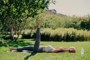 photo en plein air d'une femme en forme active fait des exercices d'aérobic avec un ballon de fitness allongé sur le karemat lève la jambe pose sur l'herbe verte avec les yeux fermés étant seul avec la nature bénéficie d'une atmosphère tranquille