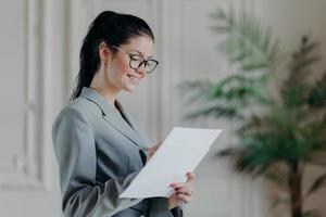 une avocate heureuse écrit dans des documents papier d'entreprise, porte des lunettes optiques, des vêtements formels, se tient dans son propre cabinet, développe une nouvelle stratégie, a une expression heureuse. personnes, carrière, concept d'emploi photo