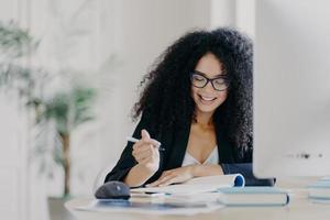 photo d'une femme aux cheveux bouclés heureuse écrit des informations, tient un stylo, a le sourire, porte des lunettes optiques et des vêtements formels pose sur le lieu de travail avec un ordinateur. l'étudiant écrit des idées pour le document de cours