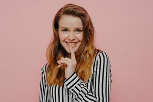 jolie jeune fille souriante regardant la caméra toucher les lèvres en se tenant debout contre le mur rose photo