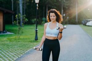 photo extérieure d'une jeune femme brune motivée porte un soutien-gorge et des leggings courts, a une silhouette mince et parfaite, soulève des haltères et entraîne des biceps, s'entraîne en plein air. concept d'exercice de remise en forme