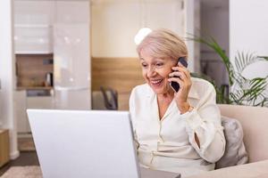 femme âgée travaillant sur un ordinateur portable, souriant, parlant au téléphone. femme âgée utilisant un ordinateur portable. femme âgée assise à la maison, utilisant un ordinateur portable et parlant sur son téléphone portable, souriante.
