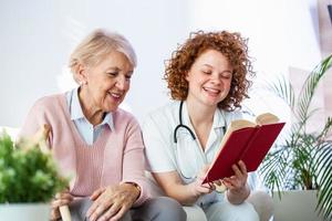 femme soignante lisant un livre assis avec une femme âgée heureuse à la maison de retraite. femme aînée heureuse assise sur un canapé blanc et écoutant une infirmière lisant un livre à haute voix