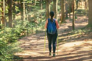 femme marchant le long d'un chemin forestier, tandis que les rayons du soleil traversent les arbres. porte un sac à dos et des bâtons de randonnée photo