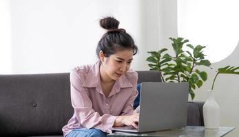 portrait d'une femme asiatique souriante assise sur le canapé, utilisant un ordinateur portable pc à l'intérieur dans le salon à la maison. espace de copie. heureuse étudiante utilisant un ordinateur pour se reposer, étudier, étudier ou travailler photo