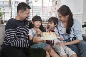 heureuse famille thaïlandaise asiatique, jeune fille est surprise avec un gâteau d'anniversaire, souffle une bougie, prie et fête joyeusement la fête avec les parents ensemble dans le salon, le style de vie de l'événement domestique de bien-être. photo