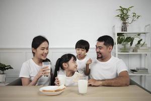 une famille thaïlandaise asiatique en bonne santé, de petits enfants et de jeunes parents boivent du lait blanc frais dans du verre et du pain joie ensemble à une table à manger le matin, bien-être nutrition maison petit déjeuner repas style de vie. photo