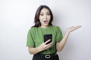 femme asiatique choquée portant un t-shirt vert pointant vers l'espace de copie à côté d'elle tenant son téléphone, isolée par fond blanc photo