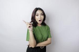 femme asiatique choquée portant un t-shirt vert pointant vers l'espace de copie à côté d'elle, isolée par fond blanc photo