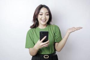 souriante belle femme asiatique pointant le doigt sur le côté vers un espace vide tout en tenant son téléphone isolé sur fond blanc photo