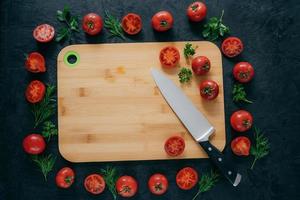 cadre de tomates autour d'une planche à découper en bois. légumes mûrs et tranches, persil vert et aneth près de la planche de cuisine et du couteau. art culinaire photo