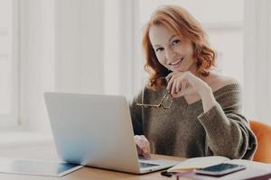 une scientifique heureuse fait des recherches sur un ordinateur portable, pose dans un espace de coworking, recherche des informations pour les travaux de cours, porte un pull chaud, se prépare pour le test de l'académie avec les technologies modernes photo