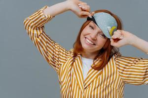 jolie jeune femme européenne a un sourire agréable, porte un bandeau sur les yeux et un pyjama rayé, se tient au-dessus du mur gris du studio, exprime de bonnes émotions, passe du bon temps pour se détendre à la maison. ambiance domestique photo