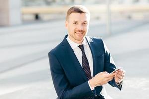 image extérieure d'un entrepreneur masculin prospère, porte un costume noir formel, une chemise blanche avec une cravate, tient un smartphone et vérifie la notification dans la rue, envoie un SMS ou vérifie la boîte e-mail en ligne photo