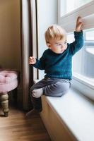 vilain petit enfant aux cheveux blonds et aux yeux bleus, assis sur le rebord de la fenêtre, regarde par la fenêtre, remarque quelque chose dans la cour. un enfant mignon et curieux joue seul, regarde dehors avec une expression attentive photo