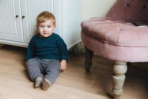 adorable bambin aux charmants yeux bleus et aux cheveux blonds, assis sur le sol contre l'intérieur de la maison, joue seul. doux petit garçon regarde directement dans la caméra, a une apparence agréable. notion d'enfance photo