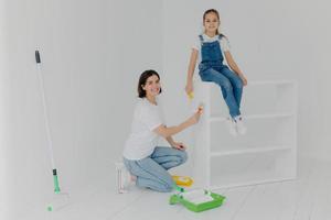 photo d'une belle mère et d'une petite fille qui travaille dur pose dans une pièce vide, peint des meubles en blanc, utilise des pinceaux, heureuse de terminer le travail, étant occupée pendant le week-end. personnes, réparation, amélioration