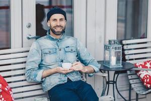 concept de personnes, de repos et de positivité. un modèle masculin barbu souriant et ravi avec une expression heureuse repose dans un café confortable, tient une tasse de thé, est de bonne humeur, pense à quelque chose d'agréable photo