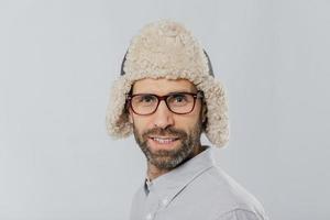 gros plan d'un homme séduisant avec chaume, porte des lunettes et un chapeau de fourrure, se prépare pour l'hiver, a des modèles de sourire doux sur fond blanc. mec masculin a les yeux bleus à poils épais se dresse à l'intérieur photo