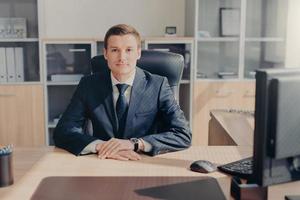 fier et beau entrepreneur masculin est assis dans son cabinet confortable, travaille sur ordinateur, satisfait d'une journée de travail réussie, habillé formellement, regarde directement dans la caméra. employé intelligent au bureau photo