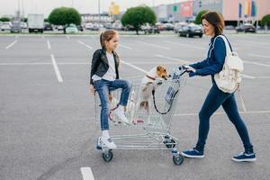 photo horizontale d'une femme heureuse porte un sac à dos, vêtue de vêtements en denim, tient un chariot, sa petite fille et deux chiens posent dans un chariot, rentrent à la maison après avoir fait du shopping dans un grand magasin. mode de vie